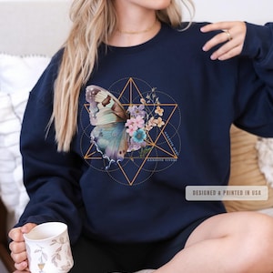 May include: Navy blue sweatshirt featuring a butterfly design with floral accents and geometric shapes. The design includes the text "Memento Vivere" and "Designed & Printed in USA". The sweatshirt is a comfortable, casual garment.