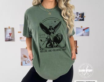 May include: A green t-shirt with a black graphic of a woman with wings standing on a lion. The text "Guided by", "WITH LOVE, I RISE - FOR JUSTICE. I FIGHT" is printed on the shirt.