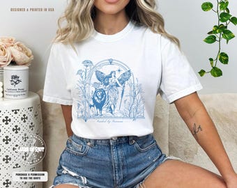 May include: White t-shirt with a blue graphic design of a woman with wings and a lion. The graphic is surrounded by a floral design and the text "Guided by Fortune".