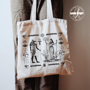 Peut inclure: Sac fourre-tout crème avec un motif noir sur le thème de l'Égypte. L'illustration comprend Anubis, une femme avec une fleur de lotus et des hiéroglyphes. Le sac a deux poignées et le texte "DESIGNED & PRINTED IN THE USA".