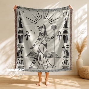 May include: A cream-colored throw blanket featuring a black and white illustration of an Egyptian goddess with cat ears, holding a staff, with pyramids in the background. The blanket has a fringed edge and a design that includes hieroglyphs and ankh symbols.