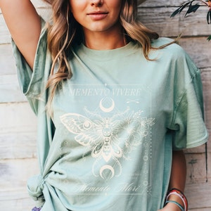 May include: A light green t-shirt with a white graphic design featuring a moth, flowers, and the text "Memento Vivere" and "Memento Mori".