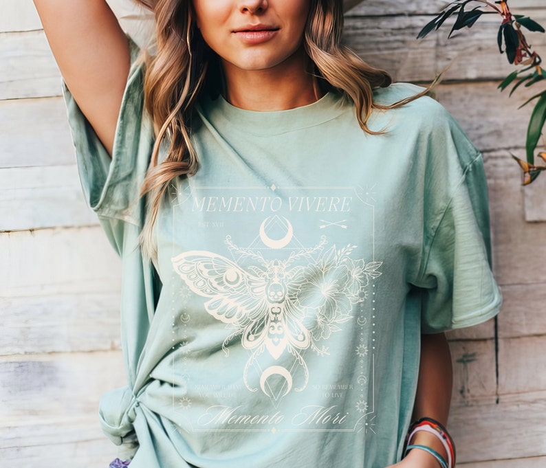 May include: A light green t-shirt with a white graphic design featuring a moth, flowers, and the text "Memento Vivere" and "Memento Mori".
