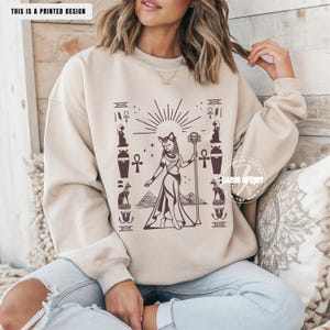 May include: Beige sweatshirt with a printed design featuring an Egyptian-themed illustration. The design includes a cat-headed figure, pyramids, and hieroglyphic symbols in brown. The text "THIS IS A PRINTED DESIGN" is at the top.