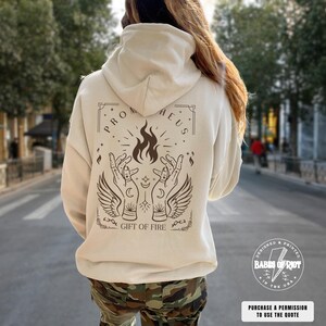 May include: A beige hoodie with a graphic on the back featuring the text "PROMETHEUS" above a flame and hands design, with the words "GIFT OF FIRE" below. The hoodie also has the text "BABES OF RIOT" and "PURCHASE A PERMISSION TO USE THE QUOTE".