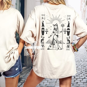 May include: Beige t-shirt with a black graphic of an Egyptian figure, pyramids, and hieroglyphs. The design features a cat-headed deity holding a staff, surrounded by sun rays and ancient symbols. The shirt has short sleeves.