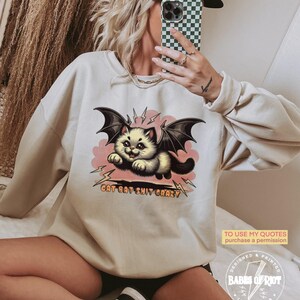May include: A light beige sweatshirt with a graphic print of a cartoon cat with bat wings flying in a pink cloud. The text "CAT BAT SHIT CRAZY" is printed below the cat.