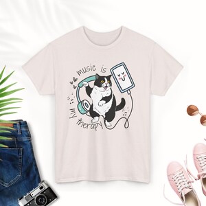 Puede incluir: Una camiseta rosa con un gato blanco y negro que lleva auriculares y escucha música en un teléfono. El texto de la camiseta dice "La música es mi terapia".