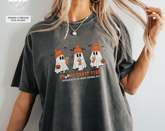 Op de afbeelding: Donkergrijs T-shirt met een afbeelding van drie cartoongeesten met oranje mutsen en drankjes en pompoenen. De tekst "Spooky Ghost Club" staat onder de geesten, met extra tekst eronder.