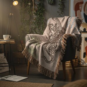 Può includere: Una coperta beige con un disegno di tigre marrone drappeggiata su una poltrona. La coperta ha un bordo con frange. La stanza ha un'atmosfera accogliente con un tavolino, una lampada e opere d'arte.