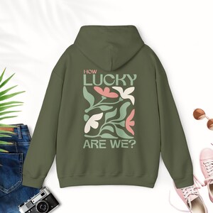 Puede incluir: Una sudadera con capucha verde con el texto "How lucky are we?" impreso en la espalda. El texto está rodeado por un diseño floral en rosa y verde.