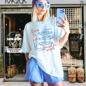 May include: White t-shirt with the text "born to fish, forced to people" and fish graphics. The shirt is paired with blue shorts. The image also shows a purple beanie, blue sunglasses, and a phone.