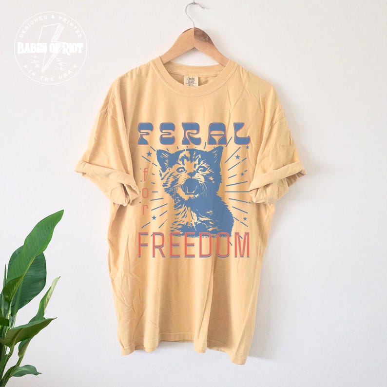 May include: Mustard yellow t-shirt with a graphic of a blue cat and the words "FERAL for FREEDOM" in blue and red. The shirt is hanging on a wooden hanger against a white background. The shirt has short sleeves and a relaxed fit.