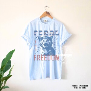 May include: Light blue t-shirt with the words "FERAL for FREEDOM" in red and blue. The graphic features a blue illustration of a cat with its mouth open. The shirt is hanging on a wooden hanger.