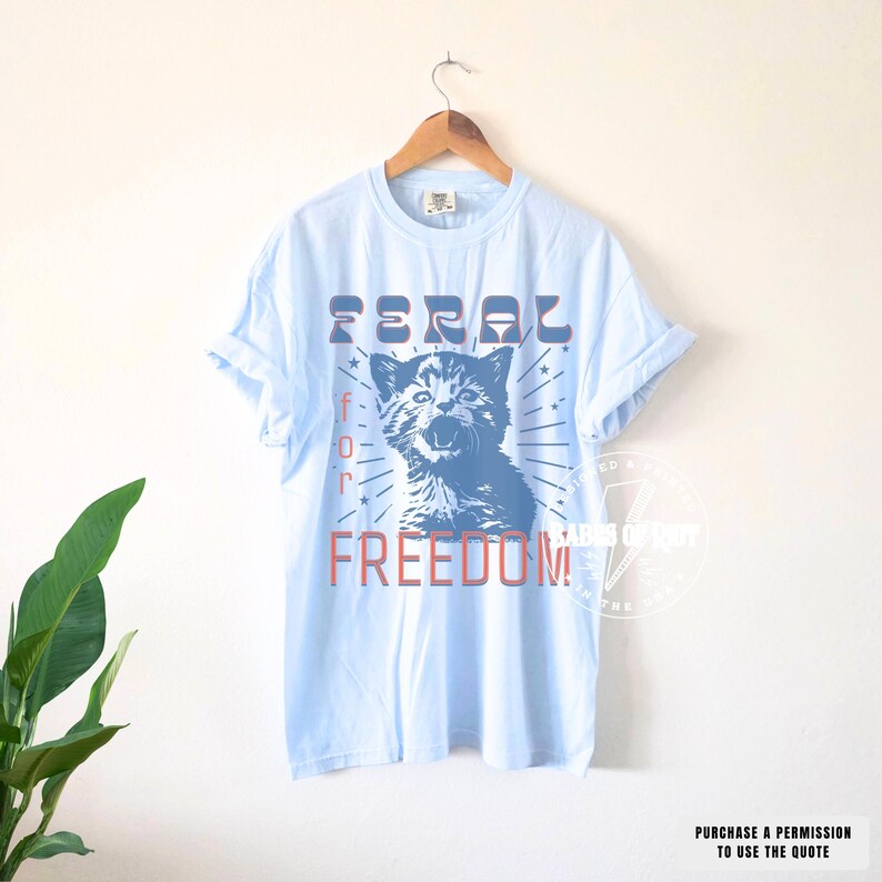 May include: Light blue t-shirt with the words "FERAL for FREEDOM" in red and blue. The graphic features a blue illustration of a cat with its mouth open. The shirt is hanging on a wooden hanger.
