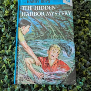 May include: A vintage hardcover book titled "The Hardy Boys: The Hidden Harbor Mystery" with a blue spine and cover art depicting two boys in a swamp, one in distress. The cover features the number 14.