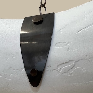 May include: A white, textured, curved surface with a black metal strap. The strap has two round, black metal rivets.