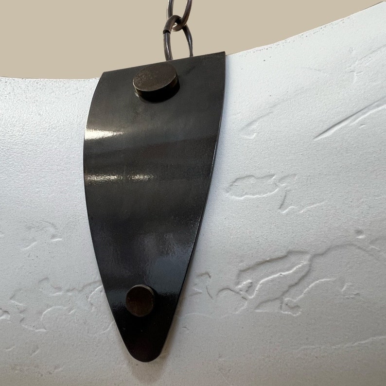 May include: A white, textured, curved surface with a black metal strap. The strap has two round, black metal rivets.