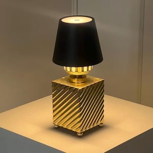 May include: A small, gold-coloured table lamp with a black lampshade. The base is a square with diagonal grooves, and the lamp has a warm glow. The lamp is approximately 30 cm tall.