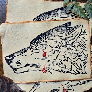 May include: A black and white print of a wolf's head on a cream-colored background. The wolf has red paint splatters on its face, giving the impression of blood.