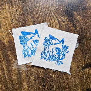 May include: Two square off-white fabric patches featuring a blue ink screen-printed design of a fox head surrounded by small flowers. The patches have raw, frayed edges and are made of a woven textile material with a visible grain and texture.