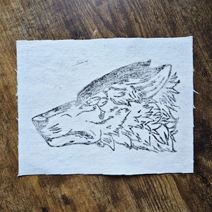 May include: A black and white drawing of a wolf's head on a rectangular piece of white fabric. The wolf is depicted in profile, with detailed fur and facial features. The fabric has a slightly rough texture.