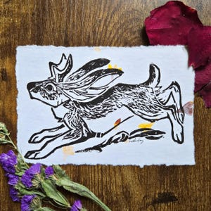 May include: Black and white linocut print of a running hare with long ears and a detailed fur pattern. The artwork is on textured white paper, with a deckled edge. Dried flowers and rose petals surround the print.