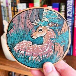 May include: A circular wooden slice featuring a hand-painted illustration of a spotted deer resting in a forest. The deer has light brown fur with white spots, and the surrounding environment includes dark teal grass and vibrant blue-green foliage.