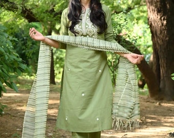 May include: A woman wearing a green salwar kameez with white embroidery and a matching dupatta. The outfit is made of a soft, flowing fabric and features a delicate floral design. The woman is standing in a garden setting.
