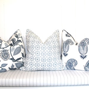May include: Three decorative throw pillows with different patterns. The pillows are on a white bench with a striped cushion. The pillows are white with blue and gray patterns. The patterns include paisley, floral, and geometric designs.