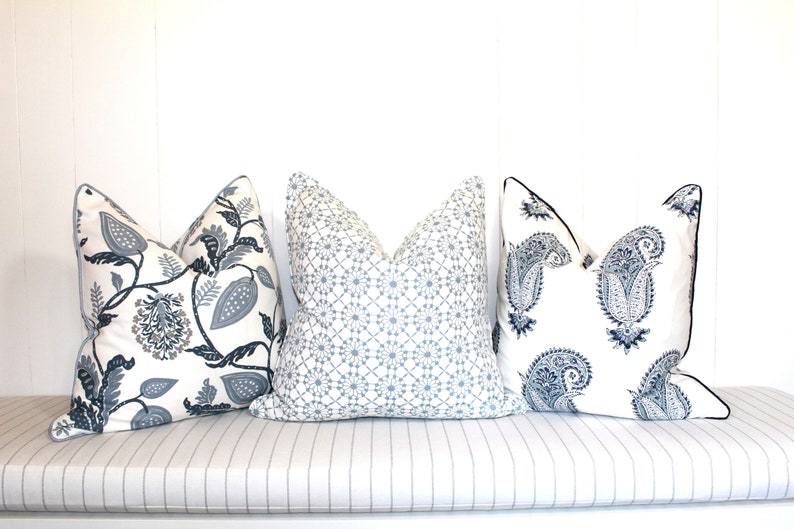 May include: Three decorative throw pillows with different patterns. The pillows are on a white bench with a striped cushion. The pillows are white with blue and gray patterns. The patterns include paisley, floral, and geometric designs.