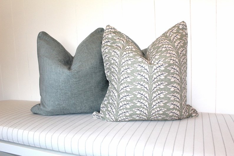 Puede incluir: Dos almohadas decorativas en un banco a rayas. Una almohada es de color verde azulado s&oacute;lido y la otra es una almohada verde estampada con un dise&ntilde;o de hojas blancas.