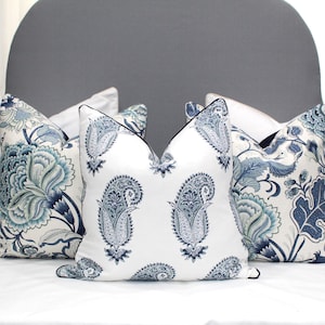 May include: Four decorative throw pillows with a white background and blue paisley and floral patterns. The pillows are arranged on a white bed with a gray headboard.