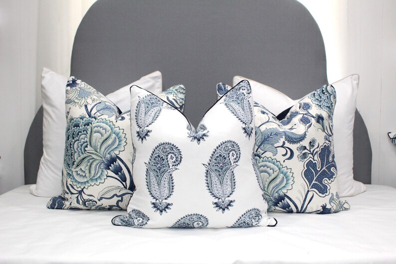 May include: Four decorative throw pillows with a white background and blue paisley and floral patterns. The pillows are arranged on a white bed with a gray headboard.