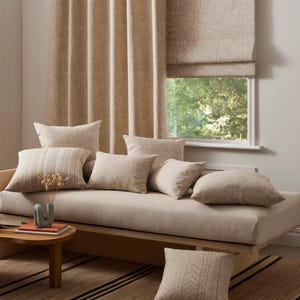 May include: A light beige sofa with a variety of throw pillows in a neutral color scheme. The sofa is positioned in front of a window with a beige patterned roman shade and a matching curtain. The sofa is sitting on a rug with black and beige stripes.