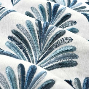May include: Close-up of a white fabric with a blue embroidered design of stylized fan-shaped leaves.