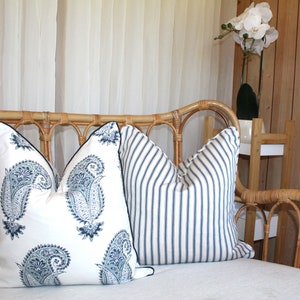May include: A white wicker sofa with two throw pillows. One pillow has a blue paisley pattern and the other has blue and white stripes. The sofa is in front of a sheer white curtain and a wooden wall.