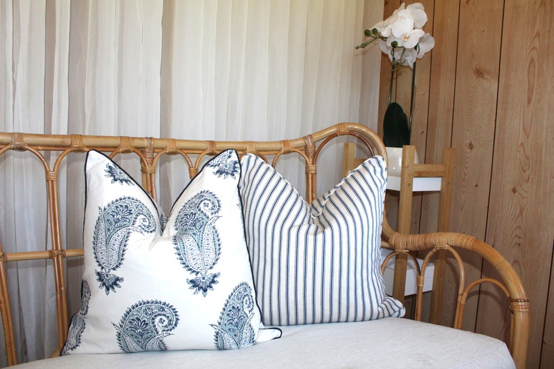 May include: A white wicker sofa with two throw pillows. One pillow has a blue paisley pattern and the other has blue and white stripes. The sofa is in front of a sheer white curtain and a wooden wall.