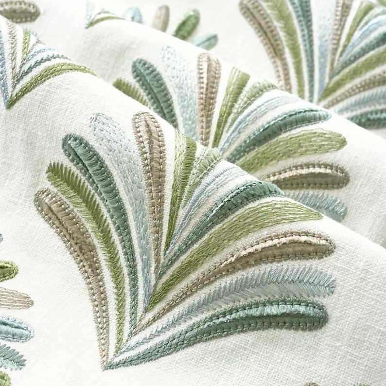 May include: Close-up of a white fabric with an embroidered design of green, blue, and brown leaves. The leaves are arranged in a fan shape and create a delicate and intricate pattern.