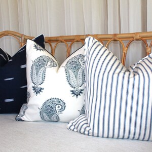 May include: Three decorative throw pillows on a light-colored couch. The pillows are made of white fabric with different patterns in shades of blue. The pillow on the left has a simple, horizontal stripe pattern. The middle pillow has a paisley pattern. The pillow on the right has a vertical stripe pattern.