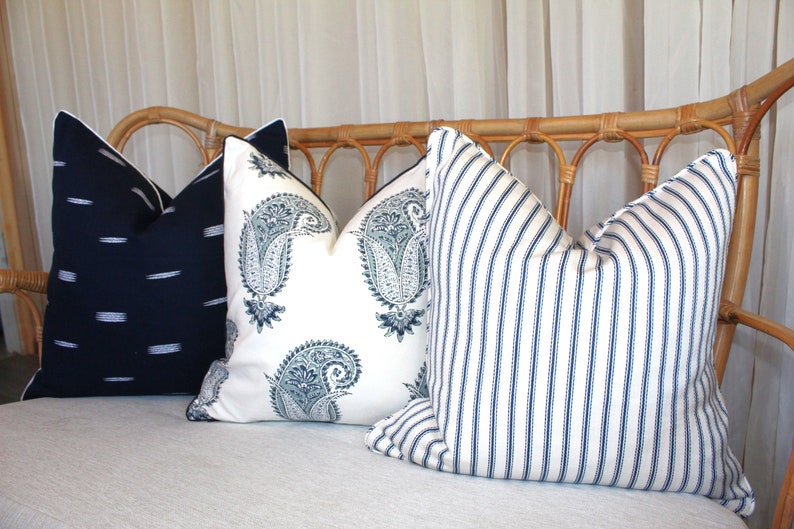 May include: Three decorative throw pillows on a light-colored couch. The pillows are made of white fabric with different patterns in shades of blue. The pillow on the left has a simple, horizontal stripe pattern. The middle pillow has a paisley pattern. The pillow on the right has a vertical stripe pattern.