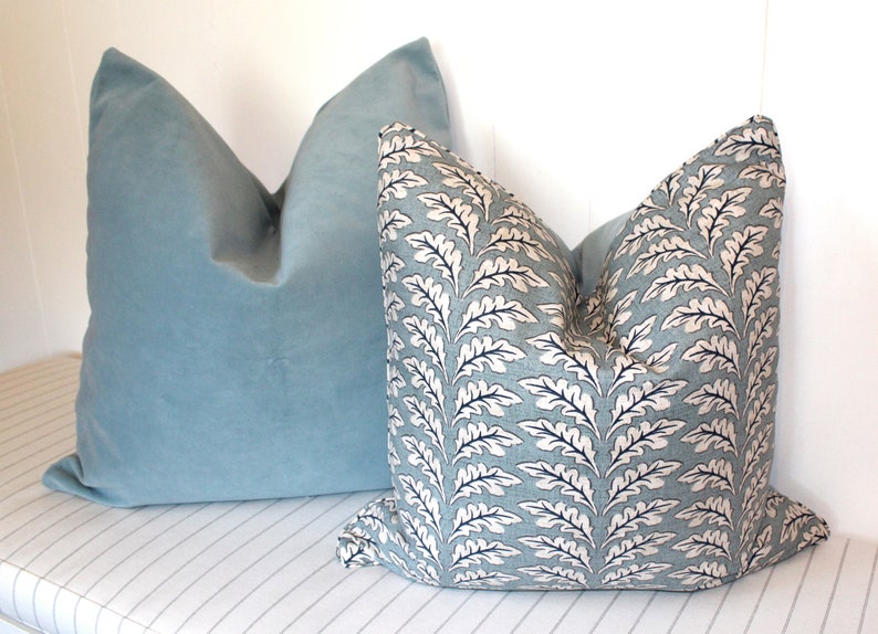 Puede incluir: Dos almohadas decorativas, una es de terciopelo azul claro liso y la otra es una almohada azul claro estampada con un dise&ntilde;o de hojas blancas y negras.