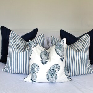 May include: A set of decorative throw pillows featuring a blue and white striped pattern and a paisley design. The pillows are arranged on a white bedspread.