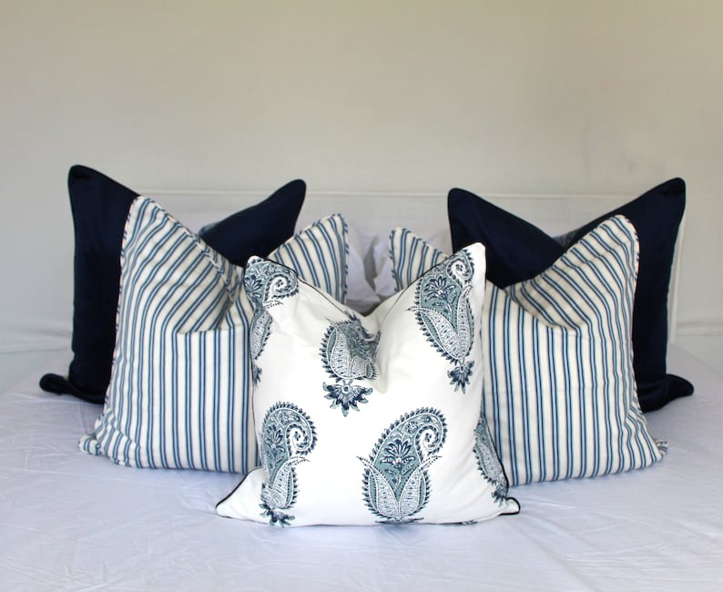 May include: A set of decorative throw pillows featuring a blue and white striped pattern and a paisley design. The pillows are arranged on a white bedspread.