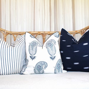 May include: Three decorative throw pillows on a white couch. The pillows are made of a white fabric with blue and white patterns. The couch is made of wicker and has a white cushion.