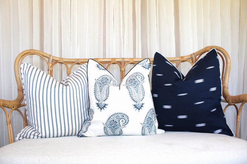 May include: Three decorative throw pillows on a white couch. The pillows are made of a white fabric with blue and white patterns. The couch is made of wicker and has a white cushion.