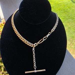 May include: A silver-toned metal necklace featuring a curb chain design with a prominent T-bar pendant drop. The necklace includes a distinct elongated oval link connector and a swivel clasp mechanism integrated into the chain structure.