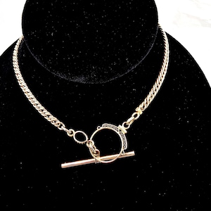 May include: A gold-toned necklace with a toggle clasp. The chain is a curb style, and the clasp features a circular design with a bar closure. The necklace is displayed on a black velvet surface.