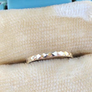 May include: A gold ring featuring a faceted, geometric surface texture that creates a series of small, reflective triangular patterns along the band. The metal has a polished finish that catches the light, highlighting the angular, repeating design.