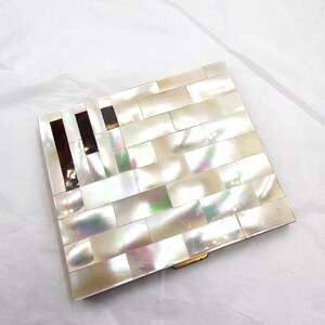 May include: A rectangular compact mirror with a mother-of-pearl mosaic design. The mosaic is made of rectangular pieces in shades of white, cream, and iridescent hues. The compact has a gold-colored metal frame and a dark brown accent on the left side.
